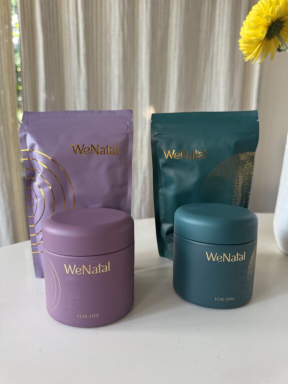 Four WeNatal branded containers, two pouches and two jars labeled "For Her" and "For Him," are displayed on a white surface with a sunflower and curtains in the background.