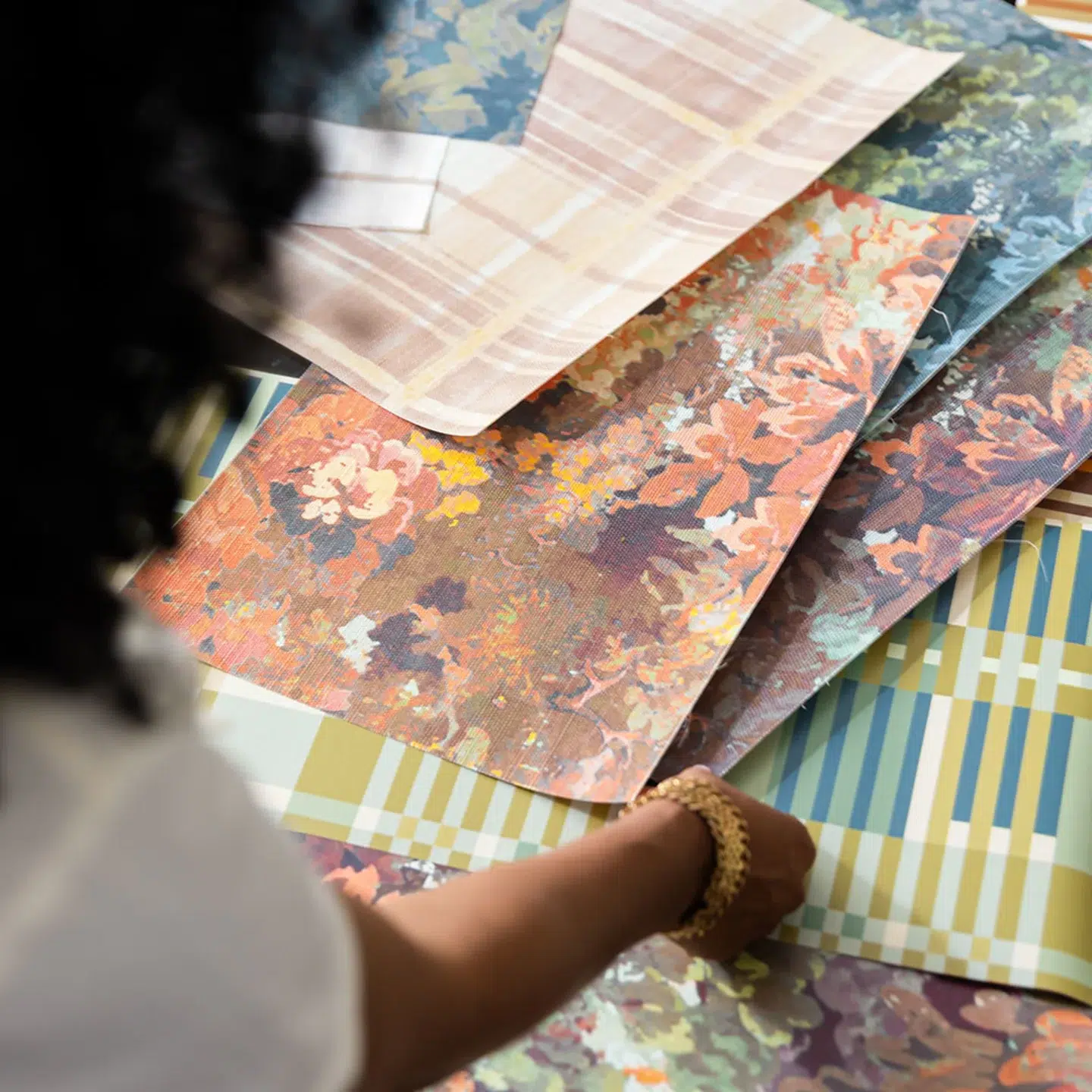 A person arranges sheets of patterned fabric samples featuring floral and plaid designs in various colors on a table.