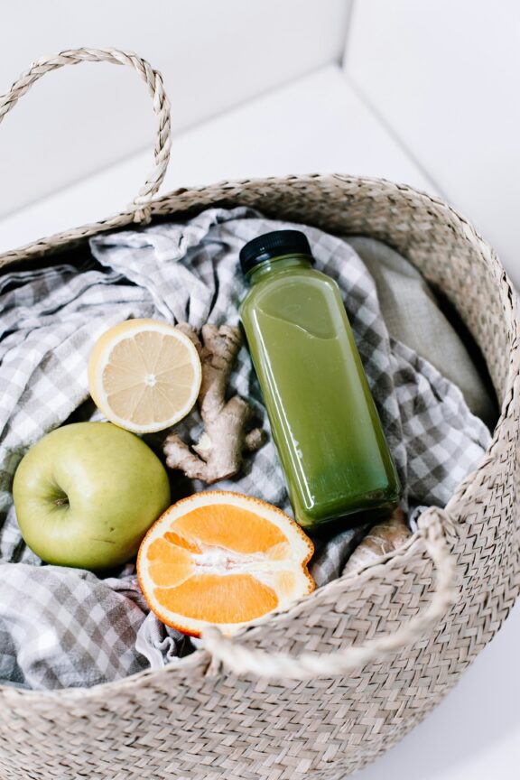 A woven basket contains a green juice bottle, a green apple, half a lemon, half an orange, ginger root, and a gray gingham cloth.