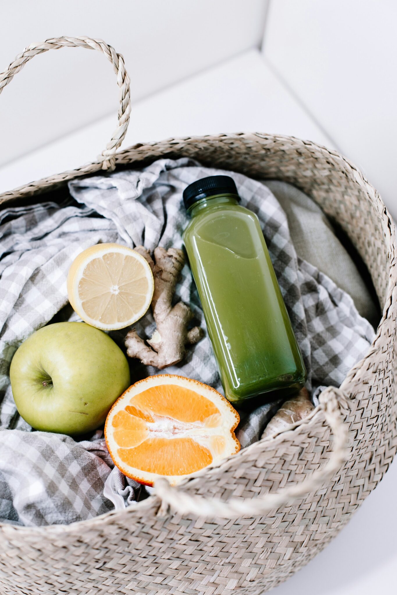 A woven basket contains a green juice bottle, a green apple, half a lemon, half an orange, ginger root, and a gray gingham cloth.