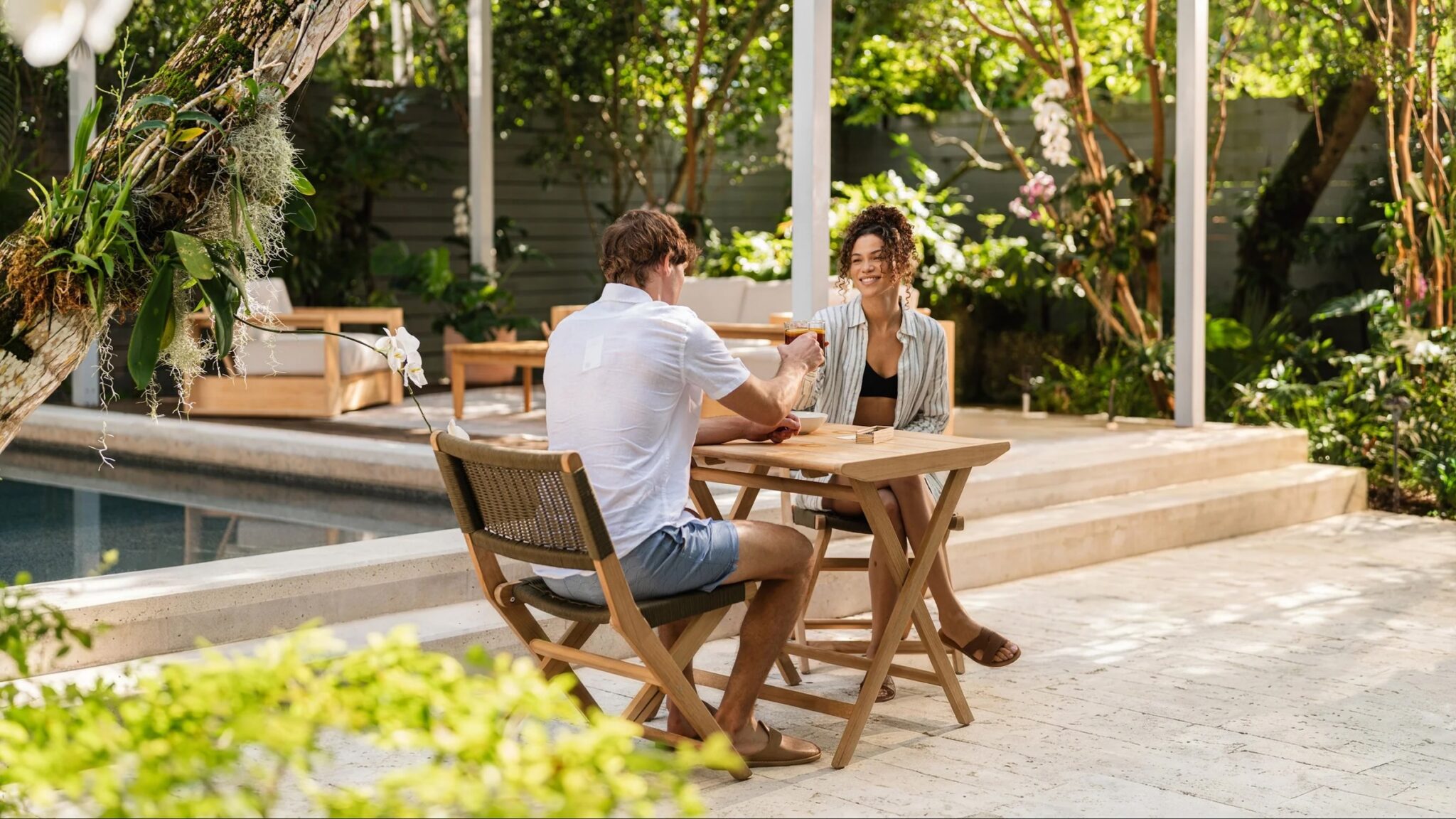 Two people sit at a wooden table outdoors by a pool, smiling and playing a game in a lush garden setting.