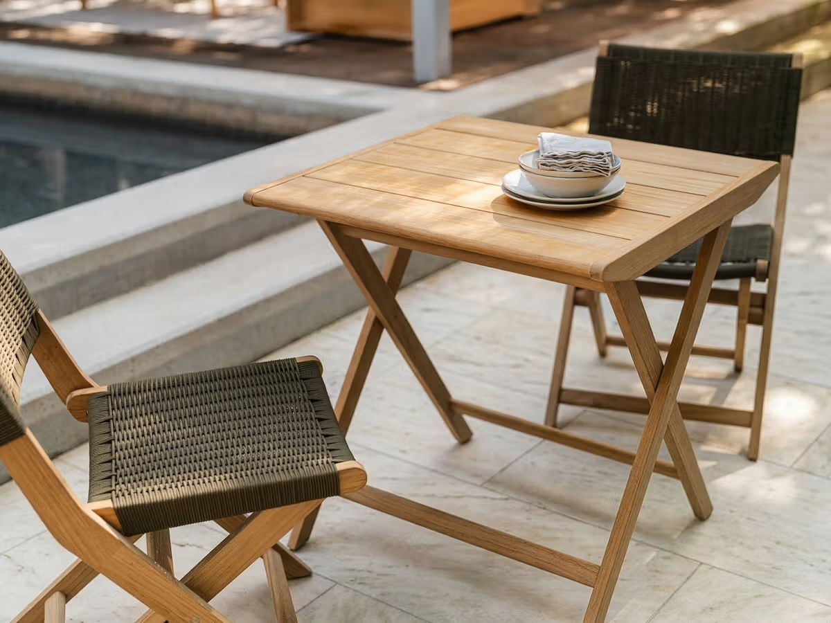 A wooden table with two woven chairs on a stone patio near a pool, set with stacked plates and napkins.