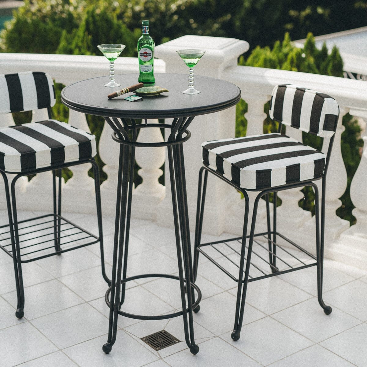 A black round table with two striped black-and-white chairs on a white tiled patio, featuring a green bottle, two cocktails, and sunglasses on the table.