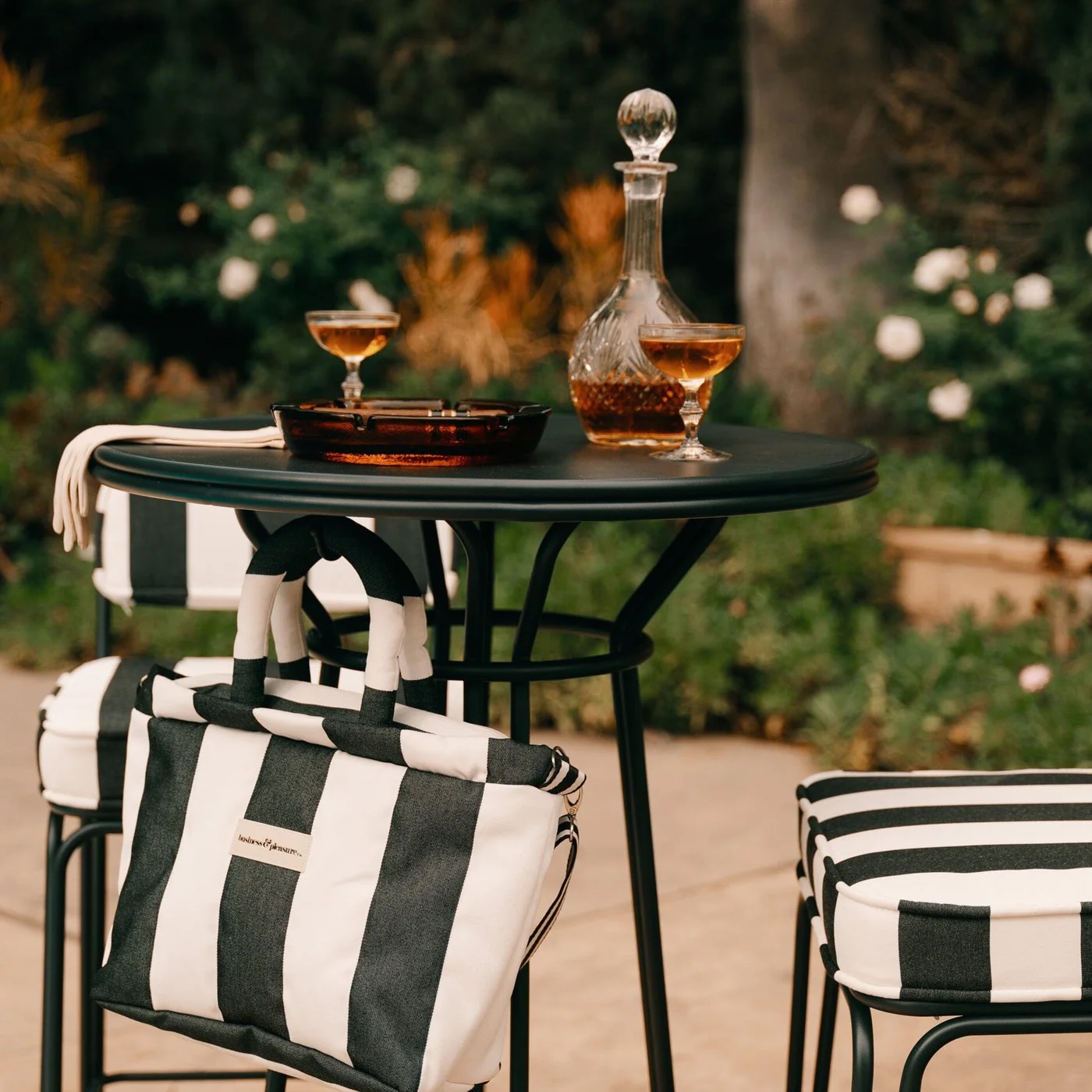 A round black table with two drinks, a glass decanter, and a dish sits next to striped cushions and a matching black-and-white striped bag outdoors.