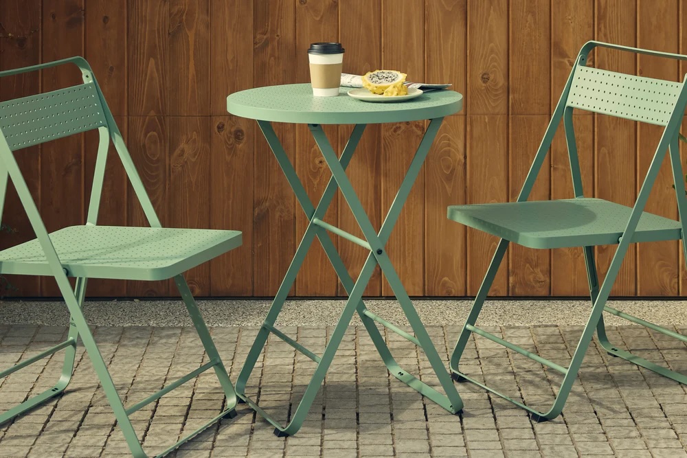 Two green metal chairs and a round table on a stone patio, with a coffee cup, a halved passion fruit, and a spoon on the tabletop, set against a wooden wall.