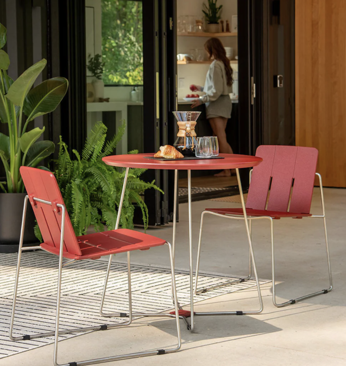 A round red table with two matching chairs is set with coffee, water, and pastries on a patio. A person stands inside the house near a kitchen counter in the background.