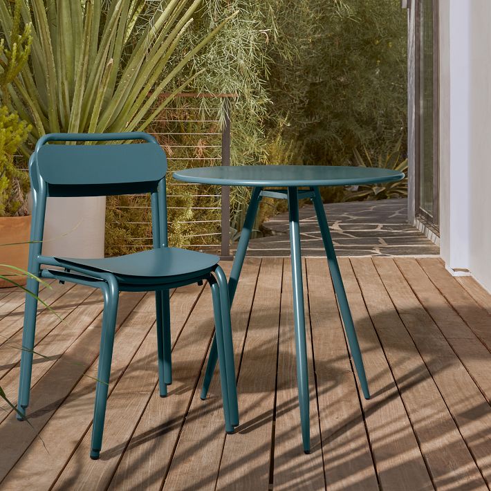 A teal metal chair and round table are placed on a wooden deck near green plants and a glass door.