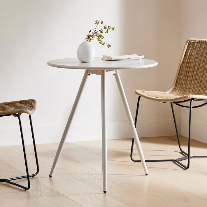 A small round white table with three legs holds a white vase with green branches and a closed book, positioned between two woven chairs on a light wood floor.