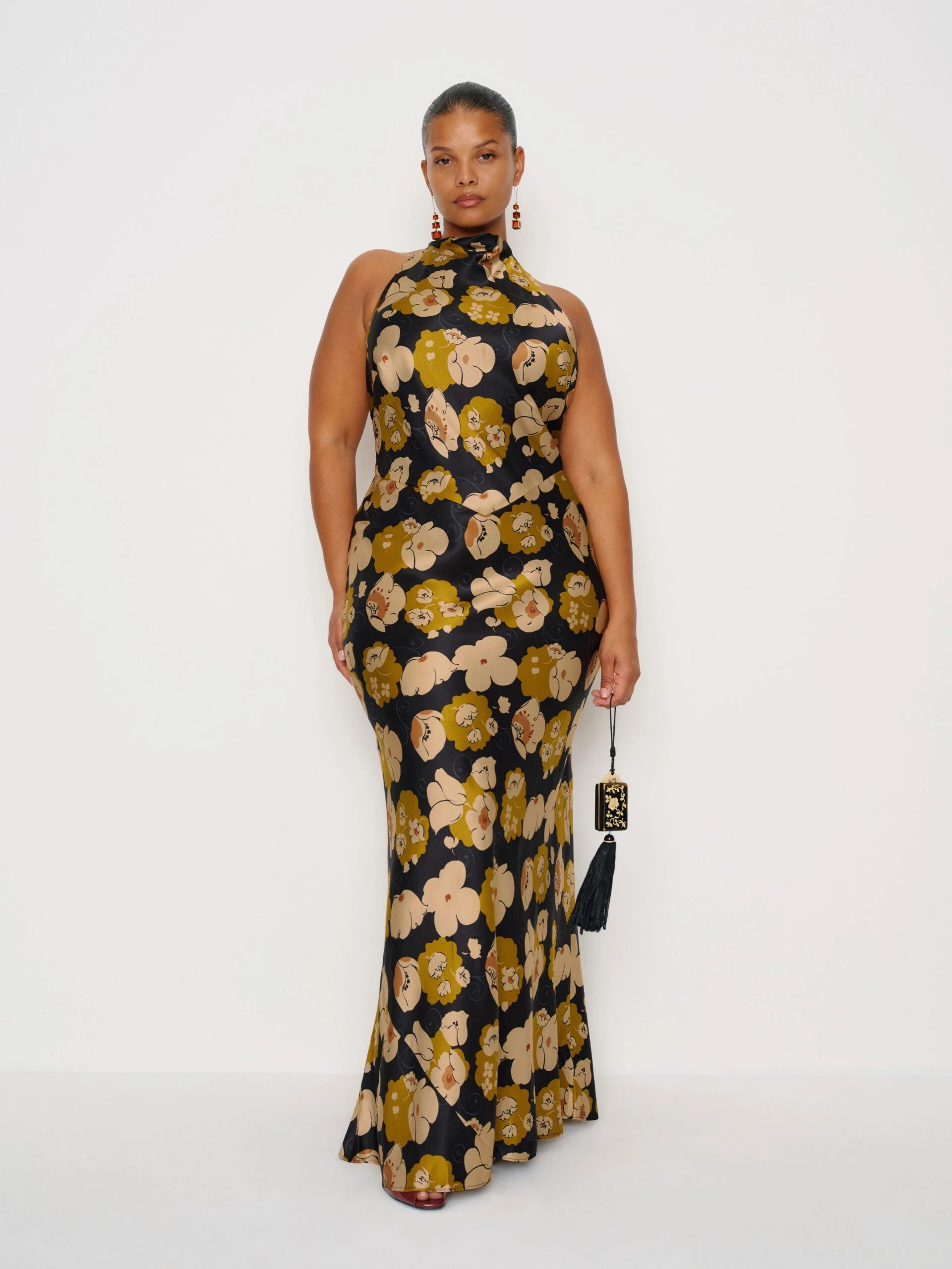 A woman poses against a white background wearing a sleeveless, floor-length black dress with a yellow and beige floral pattern, holding a small black handbag.