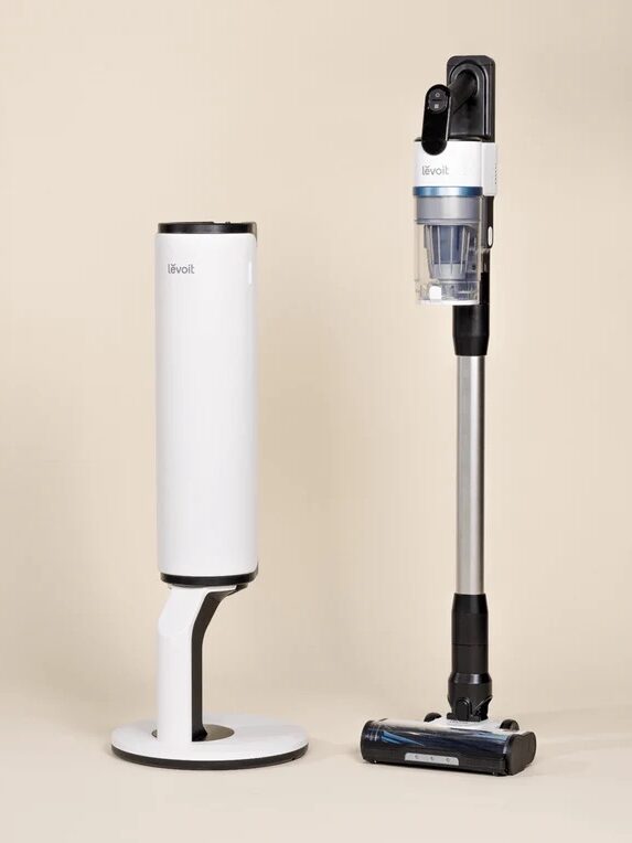 A cordless stick vacuum cleaner stands upright next to its matching white charging and storage dock, both on a neutral background.