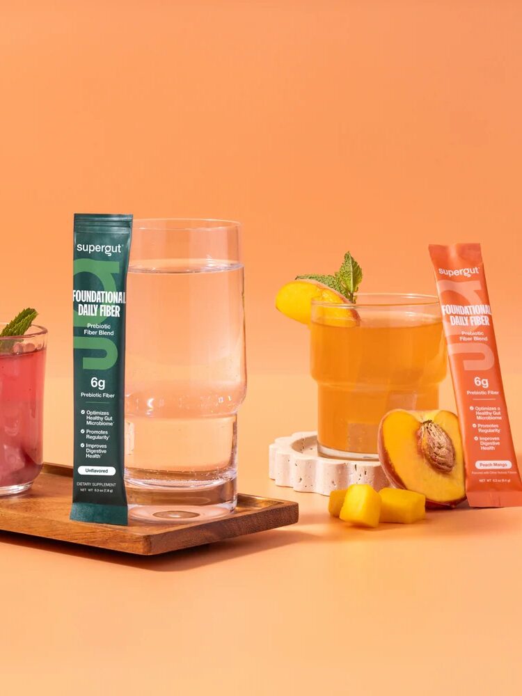 Two Supergut fiber supplements for women packets stand upright between glasses of water and fruit drinks, with fresh berries and peach slices on an orange background.