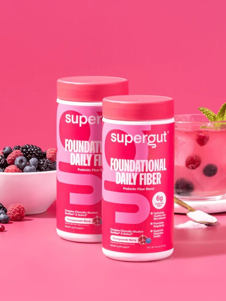Two containers of Supergut Foundational Daily Fiber powder, a top choice in fiber supplements for women, are displayed in front of a bowl of mixed berries and a glass of pink berry drink on a pink background.