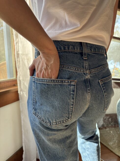 Person in a white t-shirt and blue jeans stands indoors near a window with one hand in their back pocket.