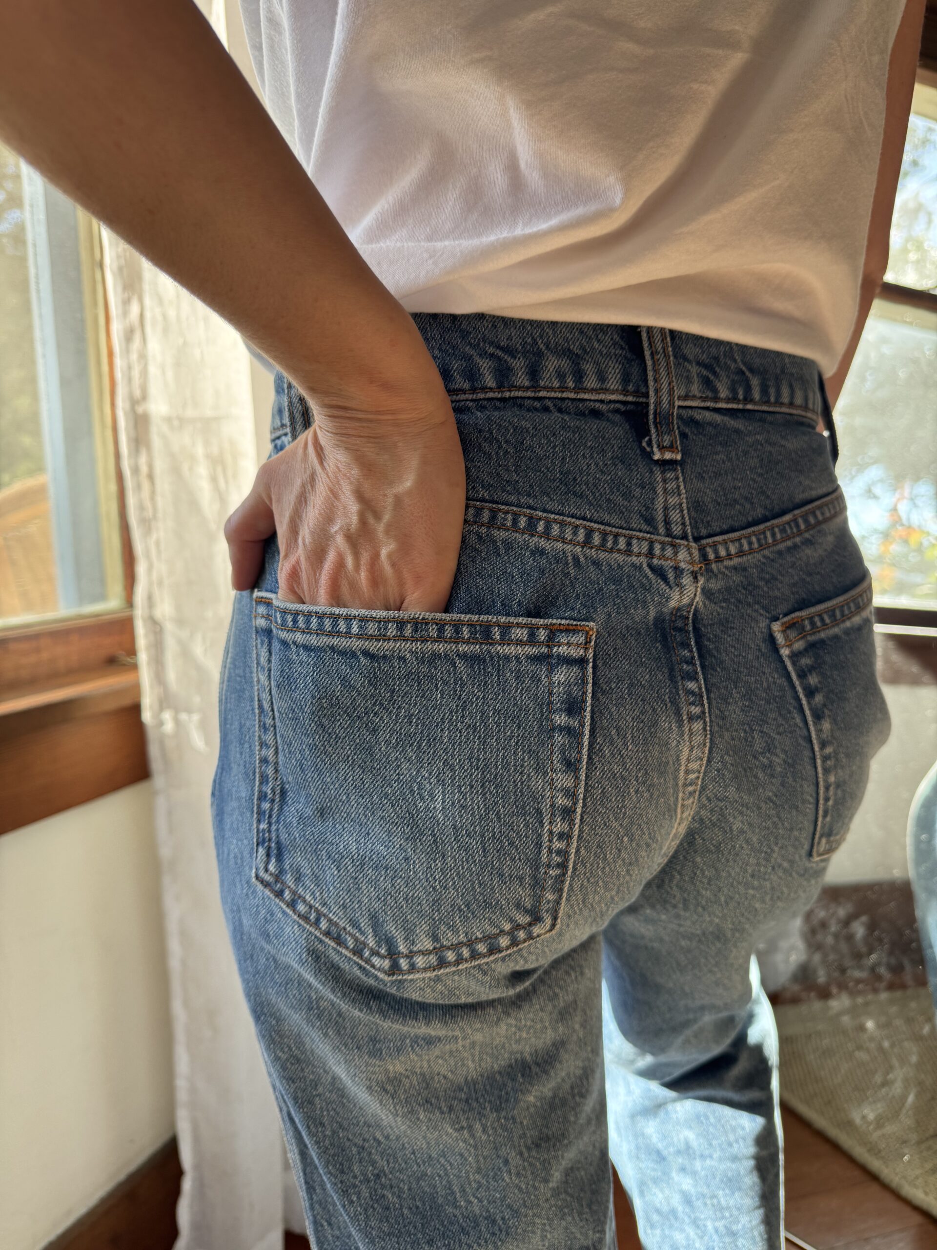 Person in a white t-shirt and blue jeans stands indoors near a window with one hand in their back pocket.