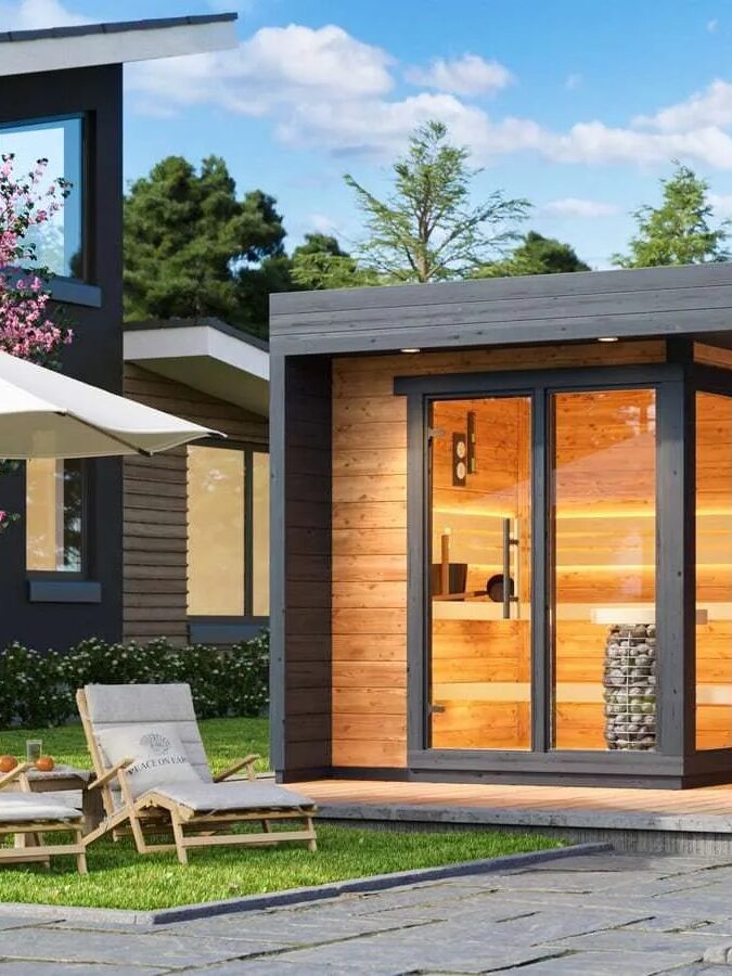 A modern wooden sauna building with large glass doors, an outdoor hot tub, two lounge chairs, and a large umbrella, surrounded by blooming trees and paved stone paths.