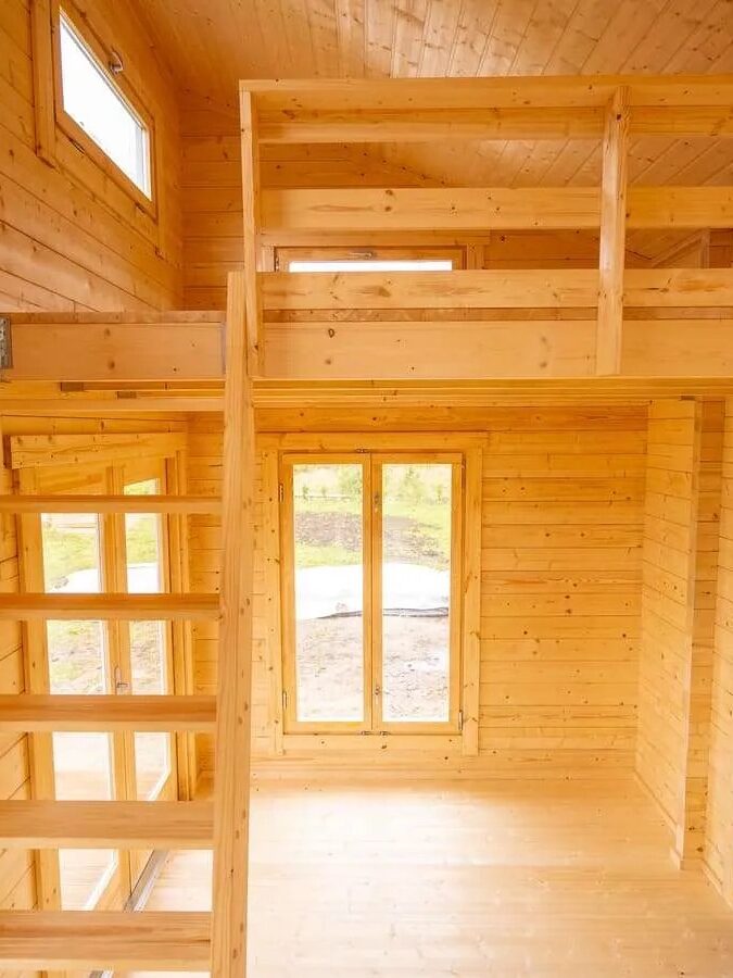 Interior of a small wooden cabin featuring a loft with railing, wooden stairs, large windows, and a black door.