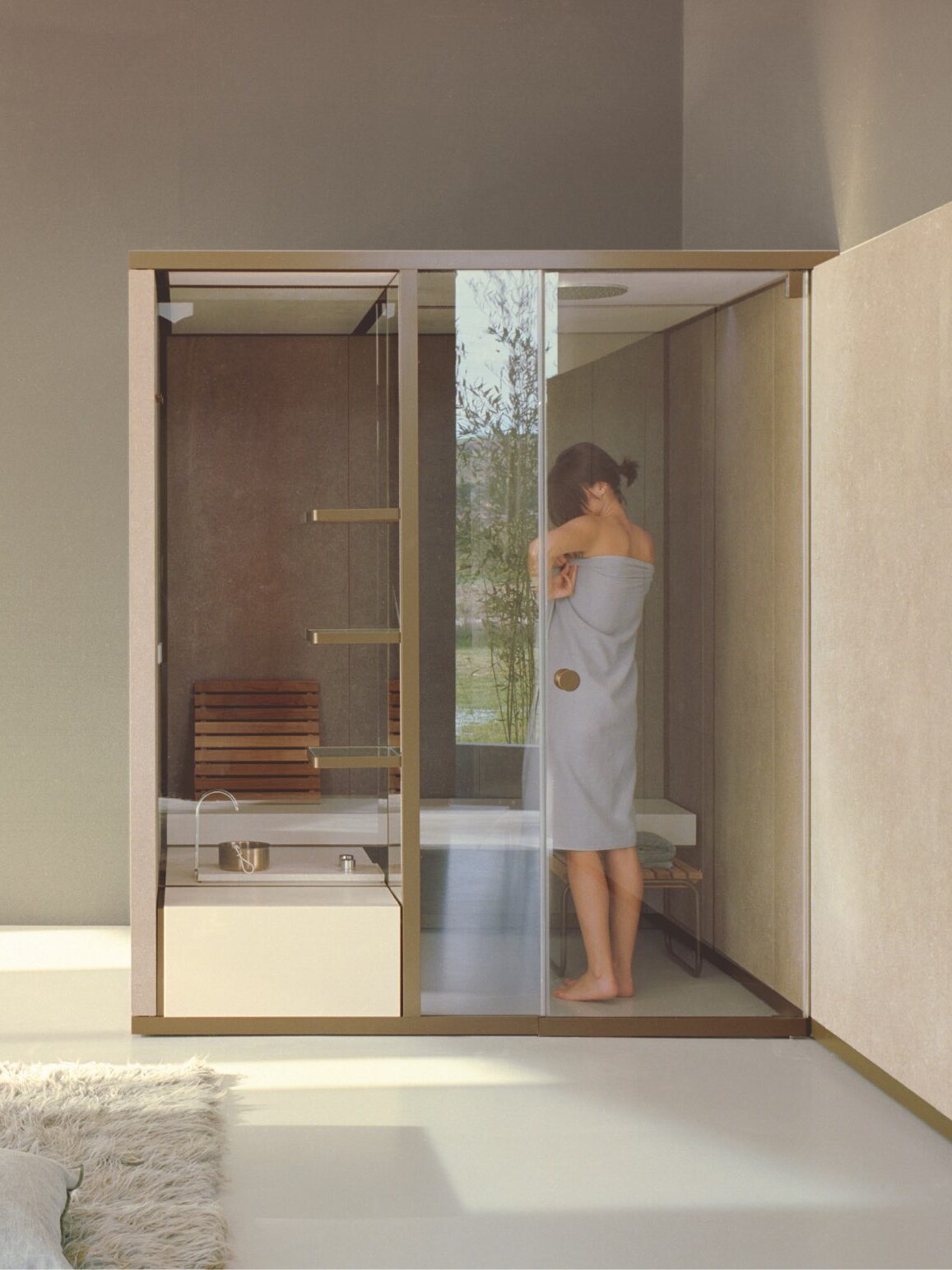 A woman wrapped in a towel stands inside a modern glass shower enclosure in a minimalist bathroom with neutral tones.