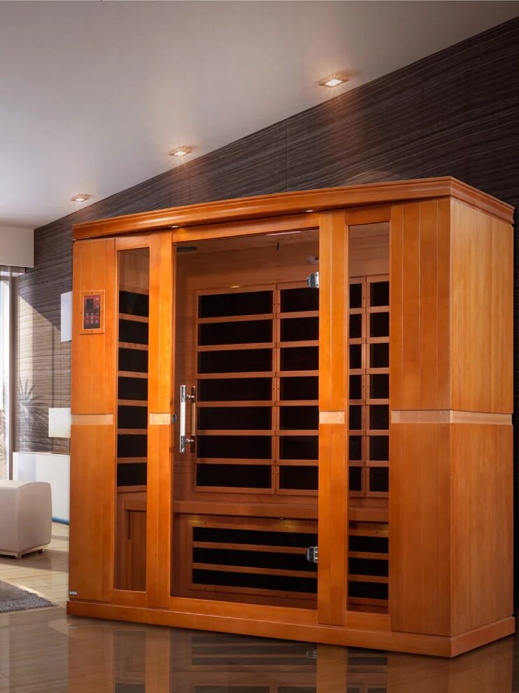 A wooden infrared sauna with glass doors is placed in the corner of a modern living room with wood panel walls and bright lighting.