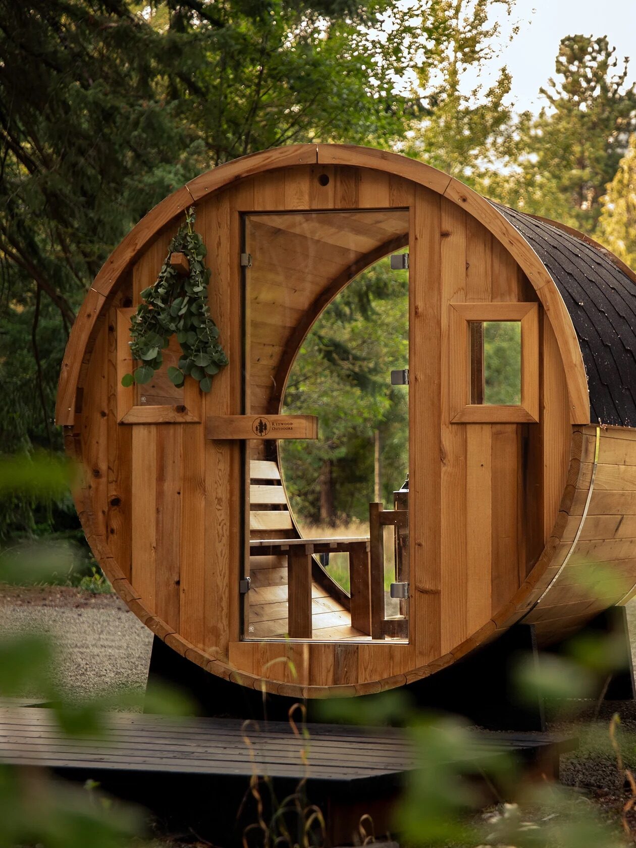A cylindrical wooden sauna with a glass door and small window is situated outdoors among trees, with a mountain visible in the background.