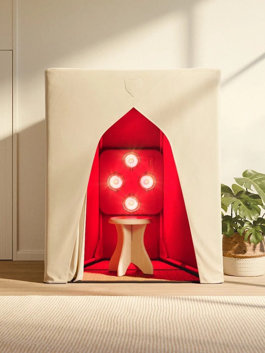 A beige tent with an open front reveals a stool and four red heat lamps inside, set in a bright room with large windows and a potted plant nearby.