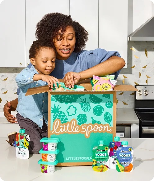 A woman and child open a Little Spoon box filled with kids’ food pouches and snacks on a kitchen counter.