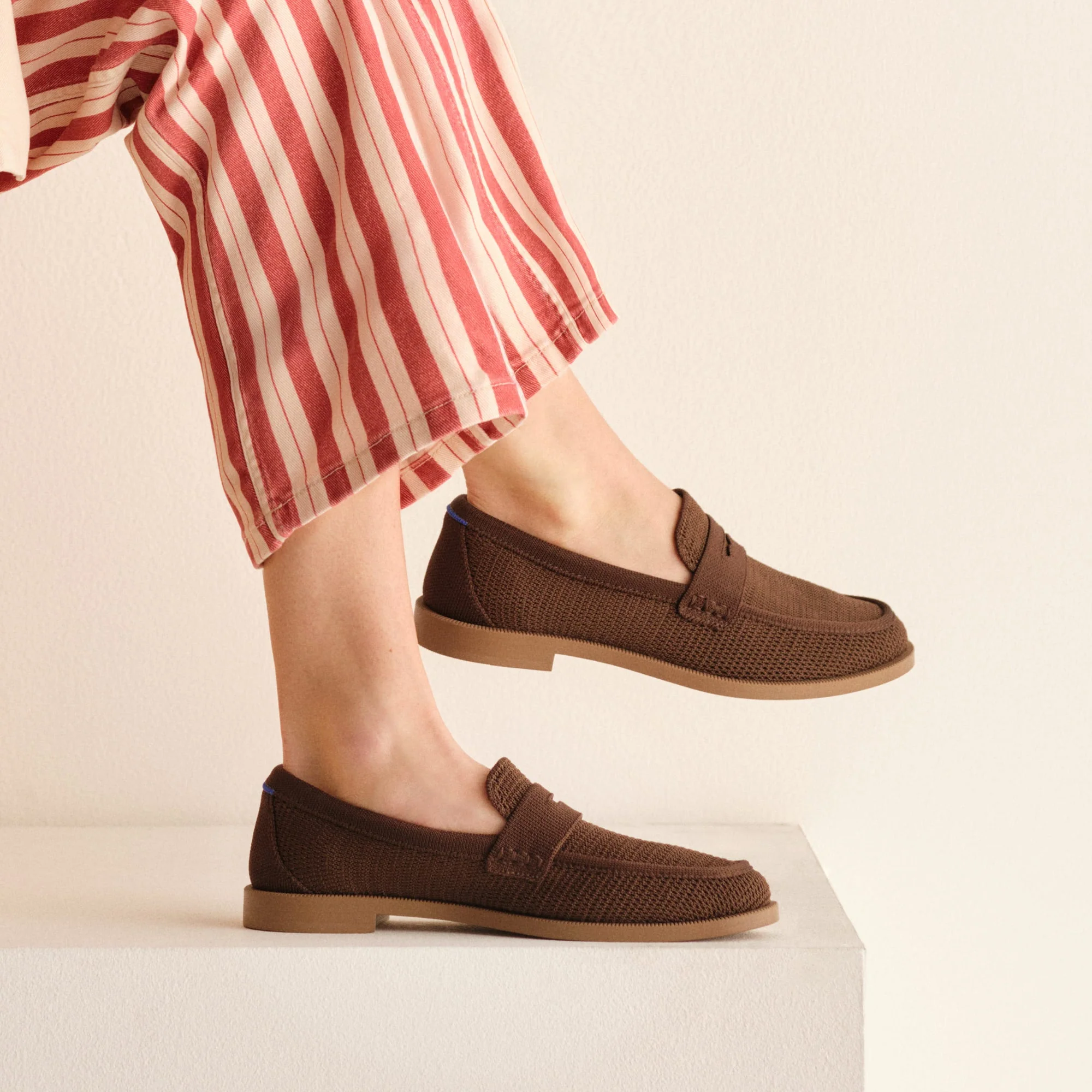 Person wearing red and white striped pants and brown loafers stands on a white platform against a plain beige background.