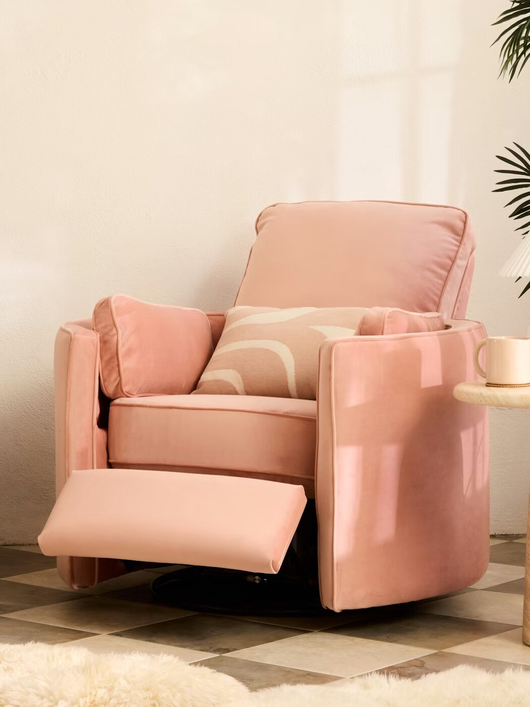 A pink recliner chair with patterned cushion, next to a round side table holding a lamp and decor, in a nursery with toys, a crib, and plants.