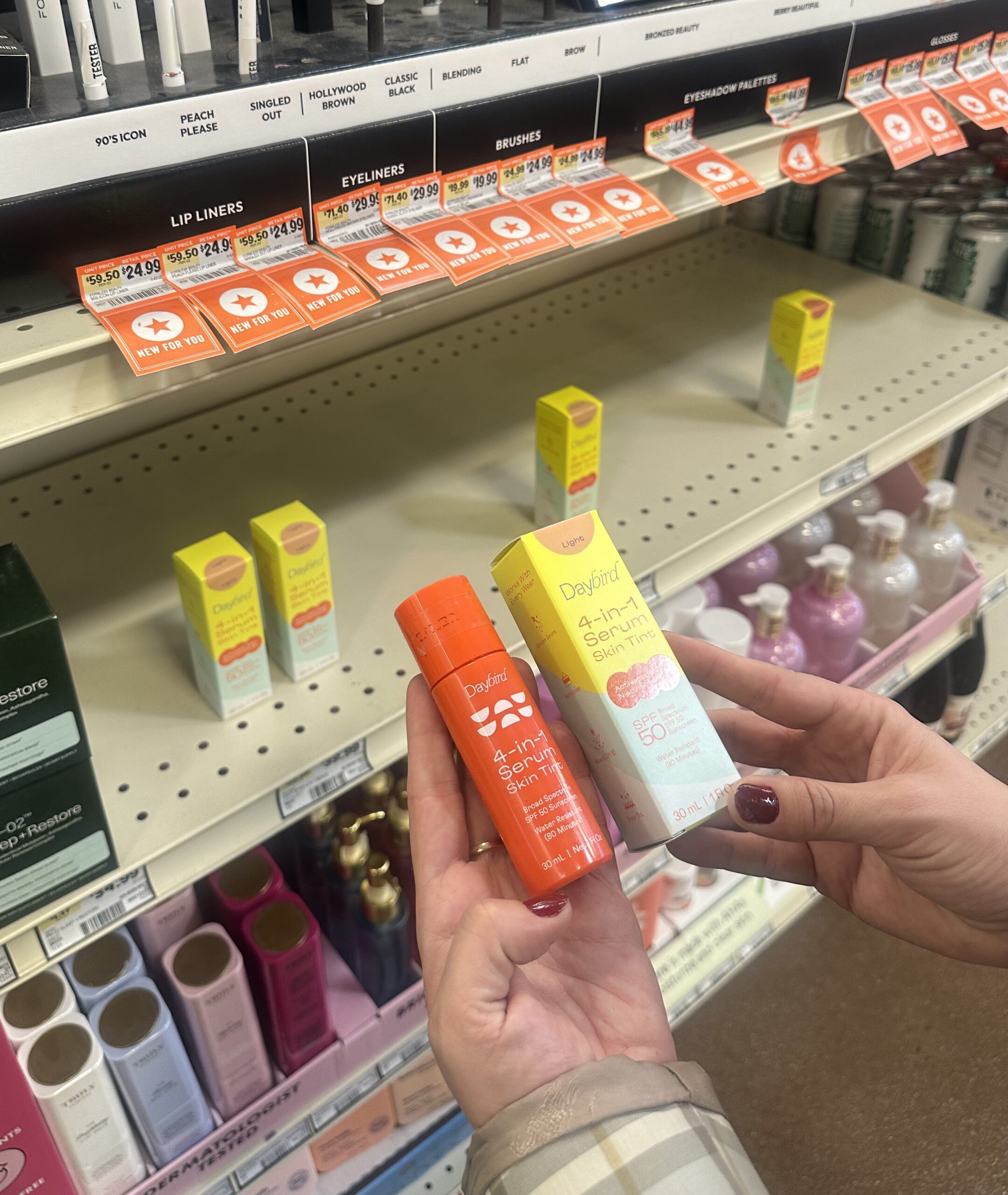 Person holding Daybird 4-in-1 Serum Skin Tint in front of a store shelf with similar skincare items. Shelves are labeled for different cosmetic products.