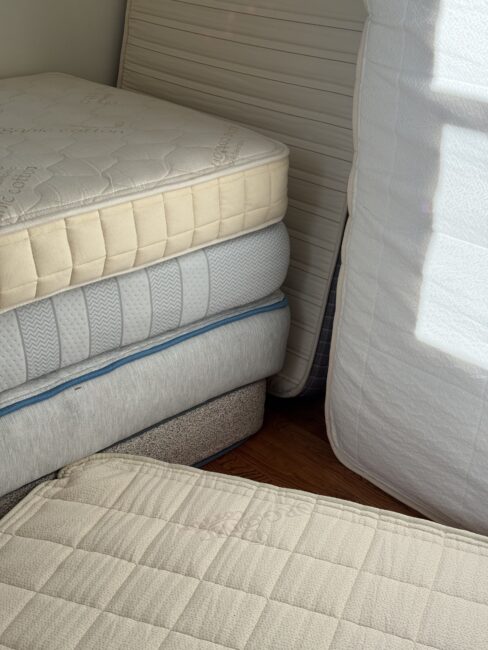 A stack of three mattresses with a fourth mattress leaning against a wall and another resting in the foreground.