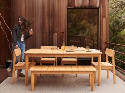 A woman holding a drink stands beside a wooden outdoor dining table set with dishes, glasses, and a bottle on a patio with wooden walls and a large window.