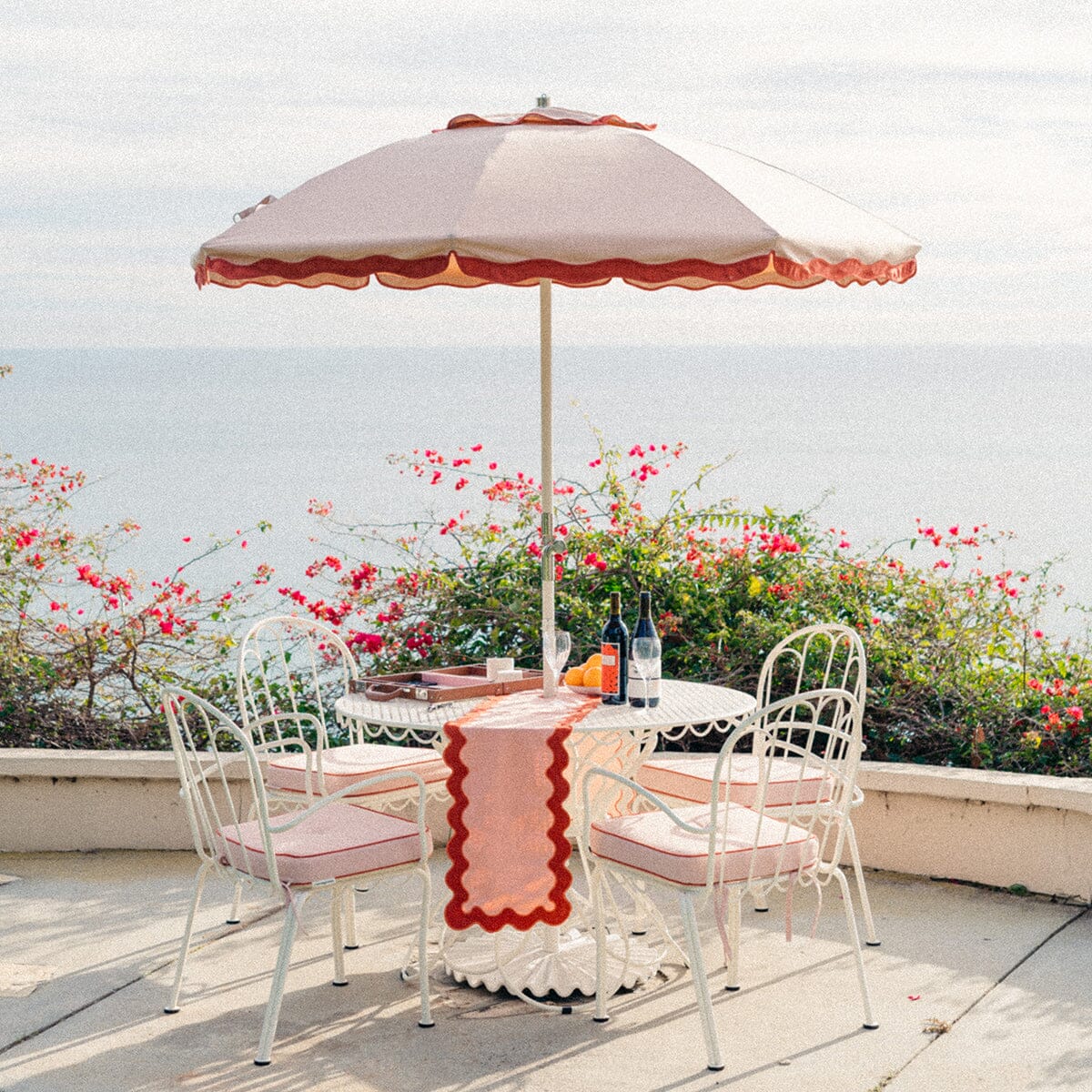 A white metal patio table with four chairs sits under a pink umbrella, set with drinks and a patterned runner, overlooking the ocean with pink flowers in the background.