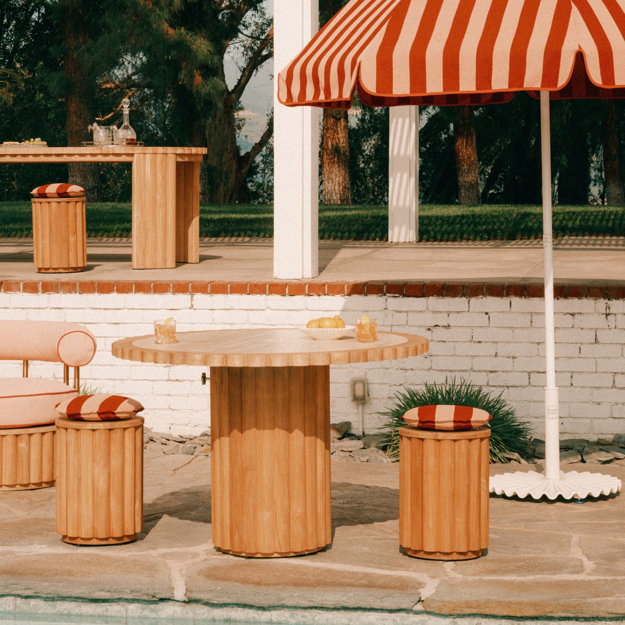 A round wooden table with three matching stools and a striped red-and-white umbrella beside a pool; drinks and food items are on the table.