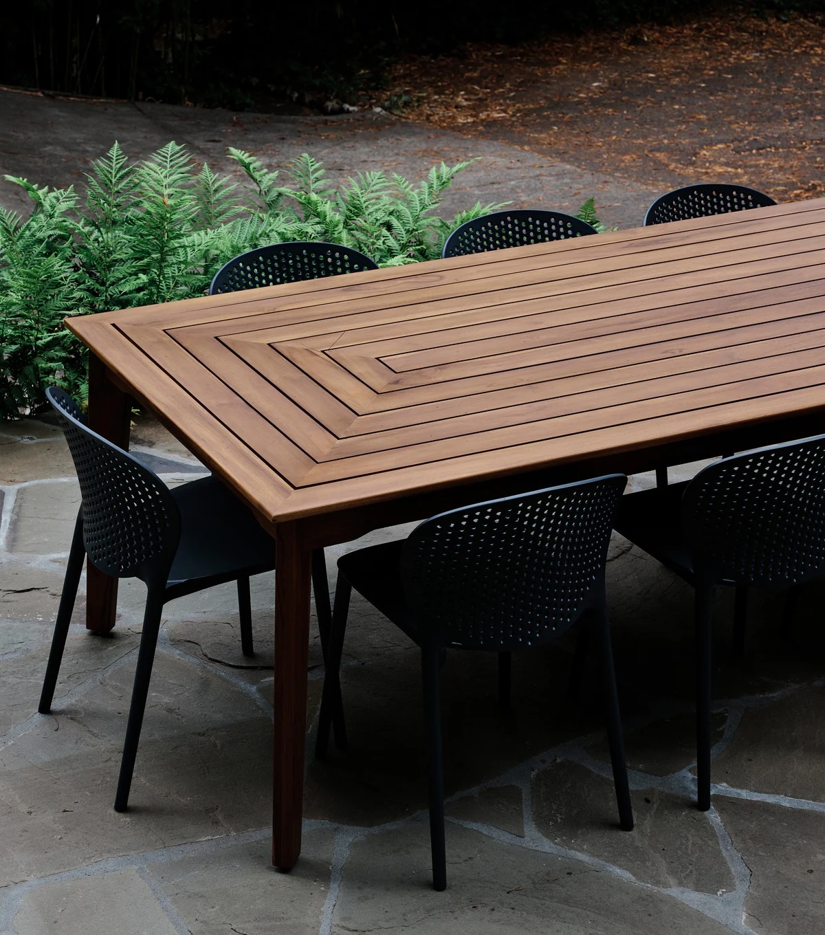 A large rectangular wooden table with a geometric design is surrounded by eight black perforated chairs on a stone patio, with green ferns in the background.