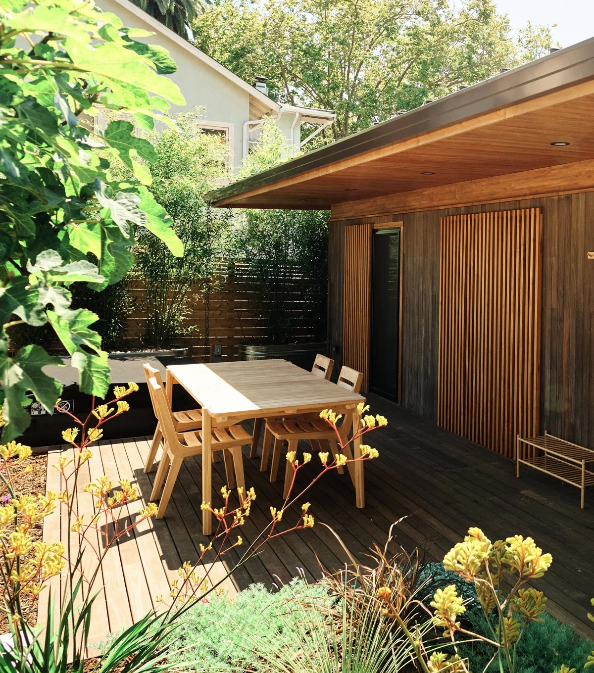A modern wooden deck with a table and four chairs, surrounded by lush greenery and plants, next to a house with vertical wooden slats.