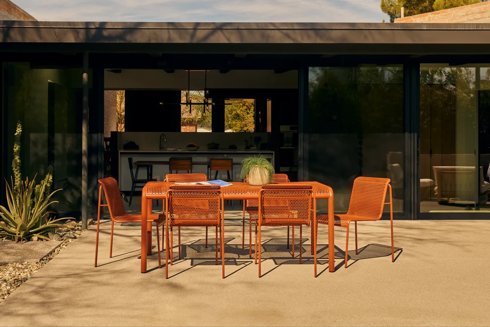 A modern outdoor dining set with orange chairs and table is placed on a patio in front of a glass-walled house. A potted plant sits on the table.
