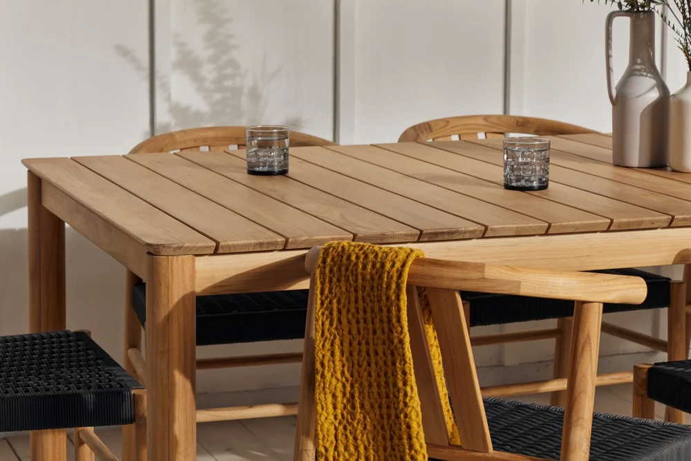 A wooden dining table with two glasses and decorative vases on top. Wooden chairs surround the table, one draped with a yellow knitted blanket.