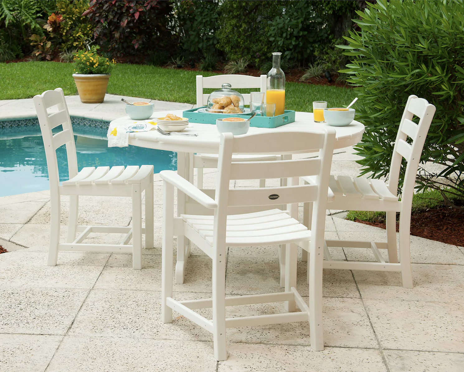 A white patio table with four chairs is set for breakfast beside a pool, featuring orange juice, pastries, and dishes on a sunny day.