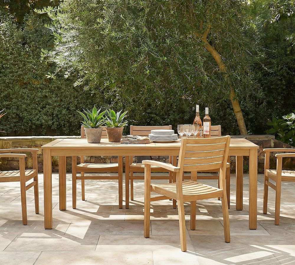 A wooden outdoor dining table with chairs, potted plants, plates, glasses, and two bottles of rosé wine set on a stone patio beneath a leafy tree.