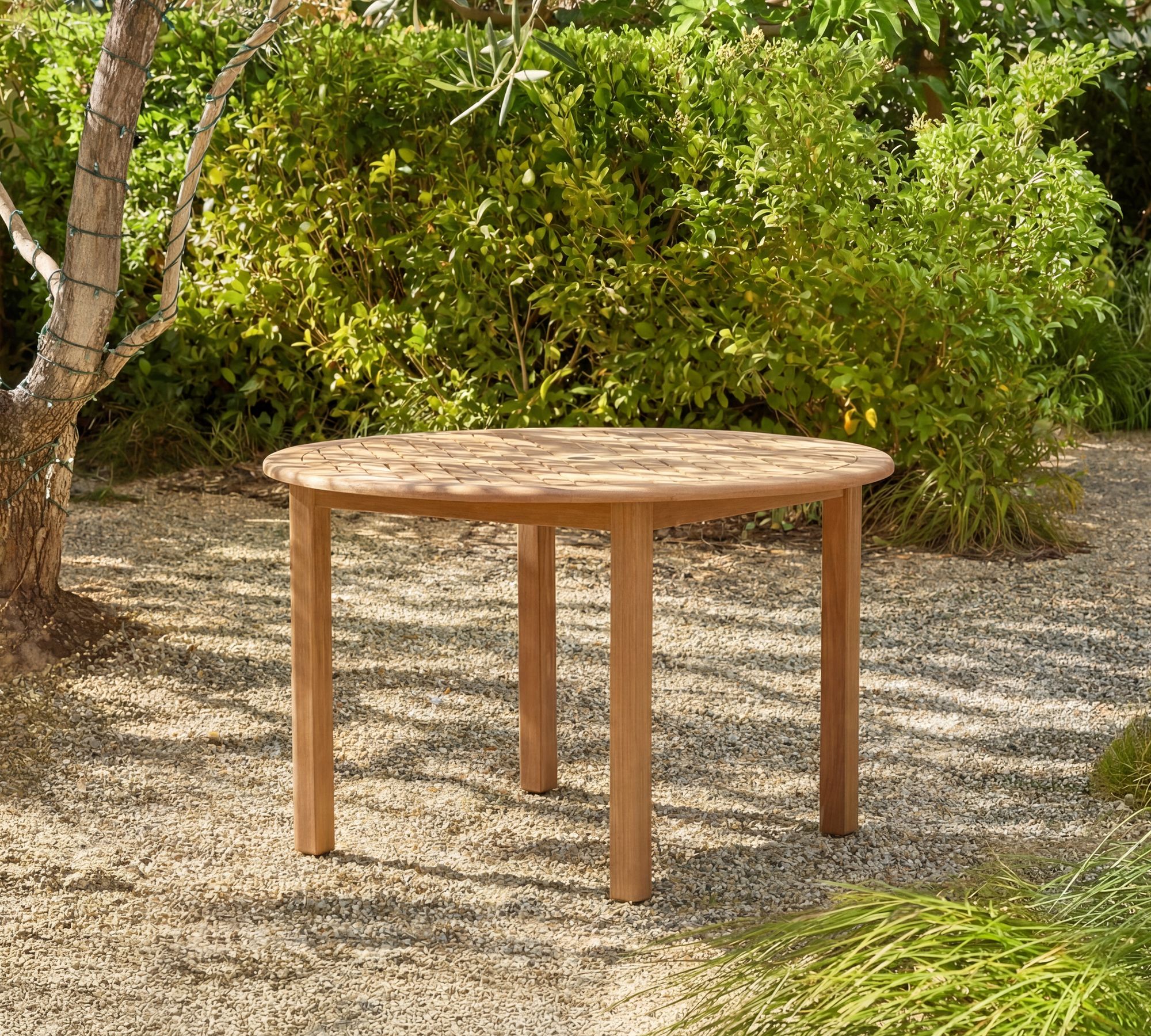 A round wooden table with four legs stands on a gravel surface outdoors, surrounded by green bushes and trees.