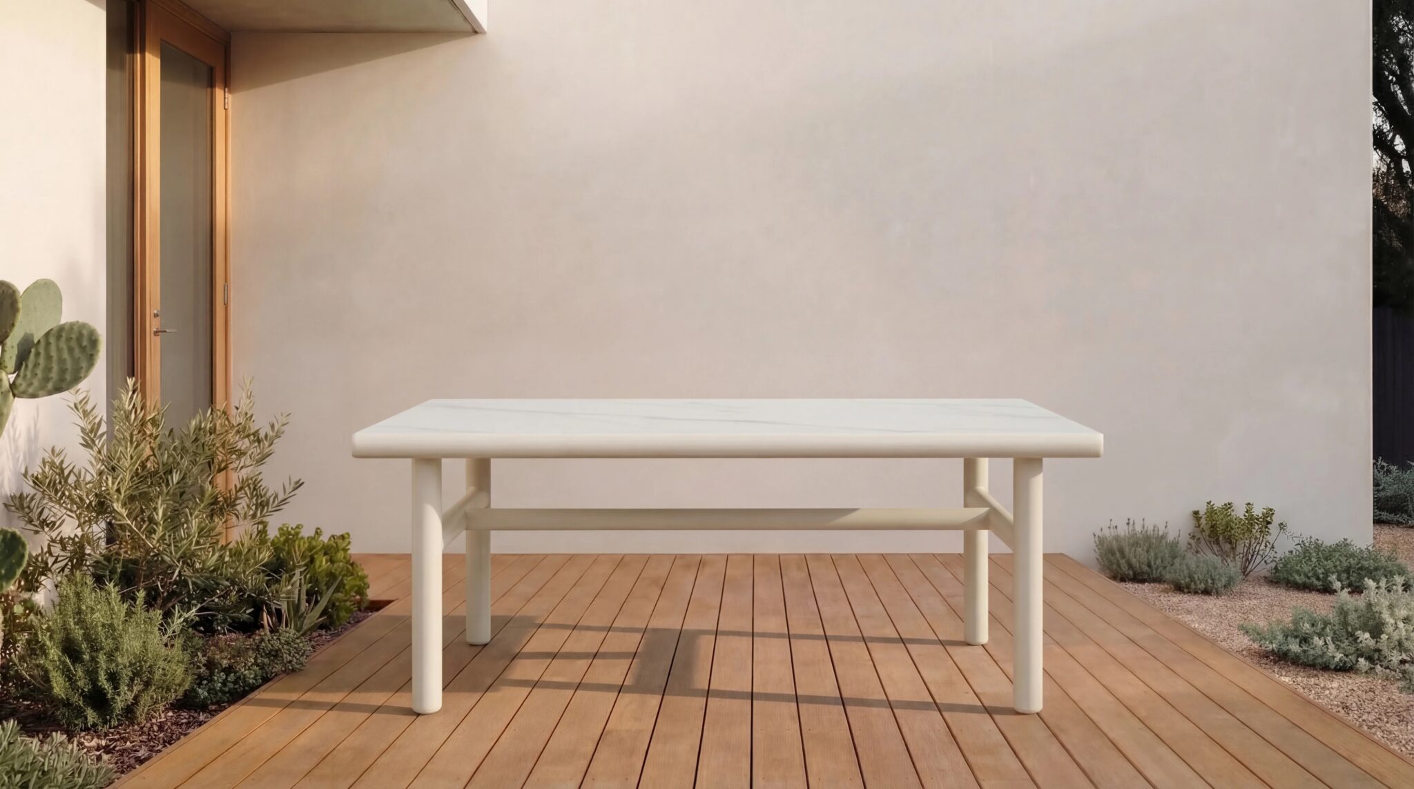 A white rectangular table stands on a wooden deck outside a modern house with light-colored walls, surrounded by plants and shrubs.