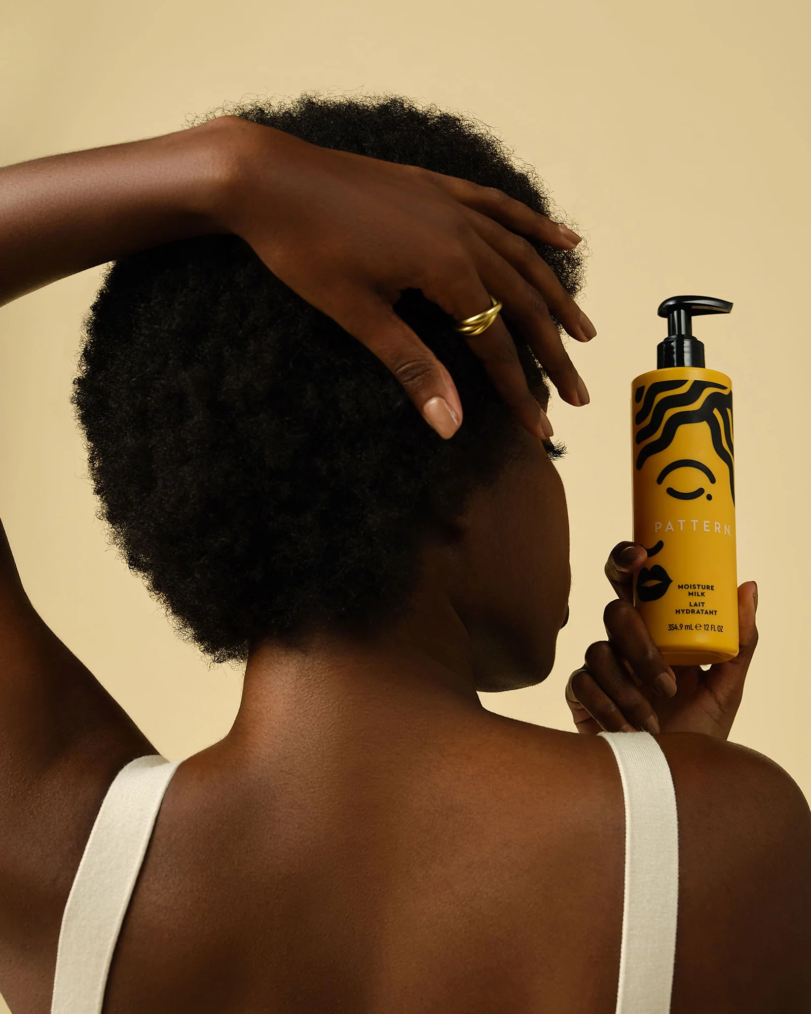 A person with short curly hair holds a yellow bottle of Pattern hair product near their head, facing away from the camera, against a beige background.