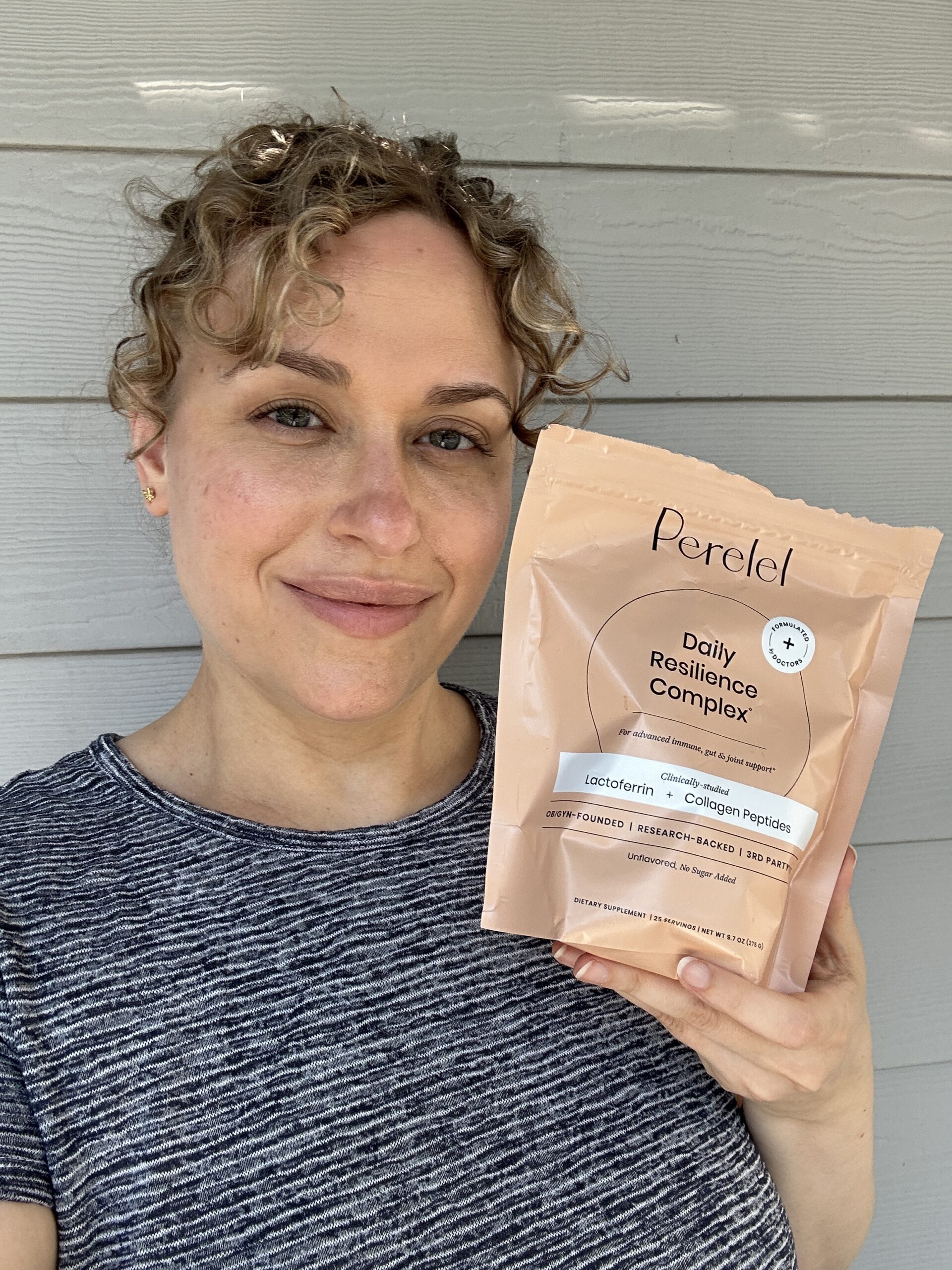 A person with curly hair holds up a beige package labeled "Perelel Daily Resilience Complex" in front of a light gray wooden wall.
