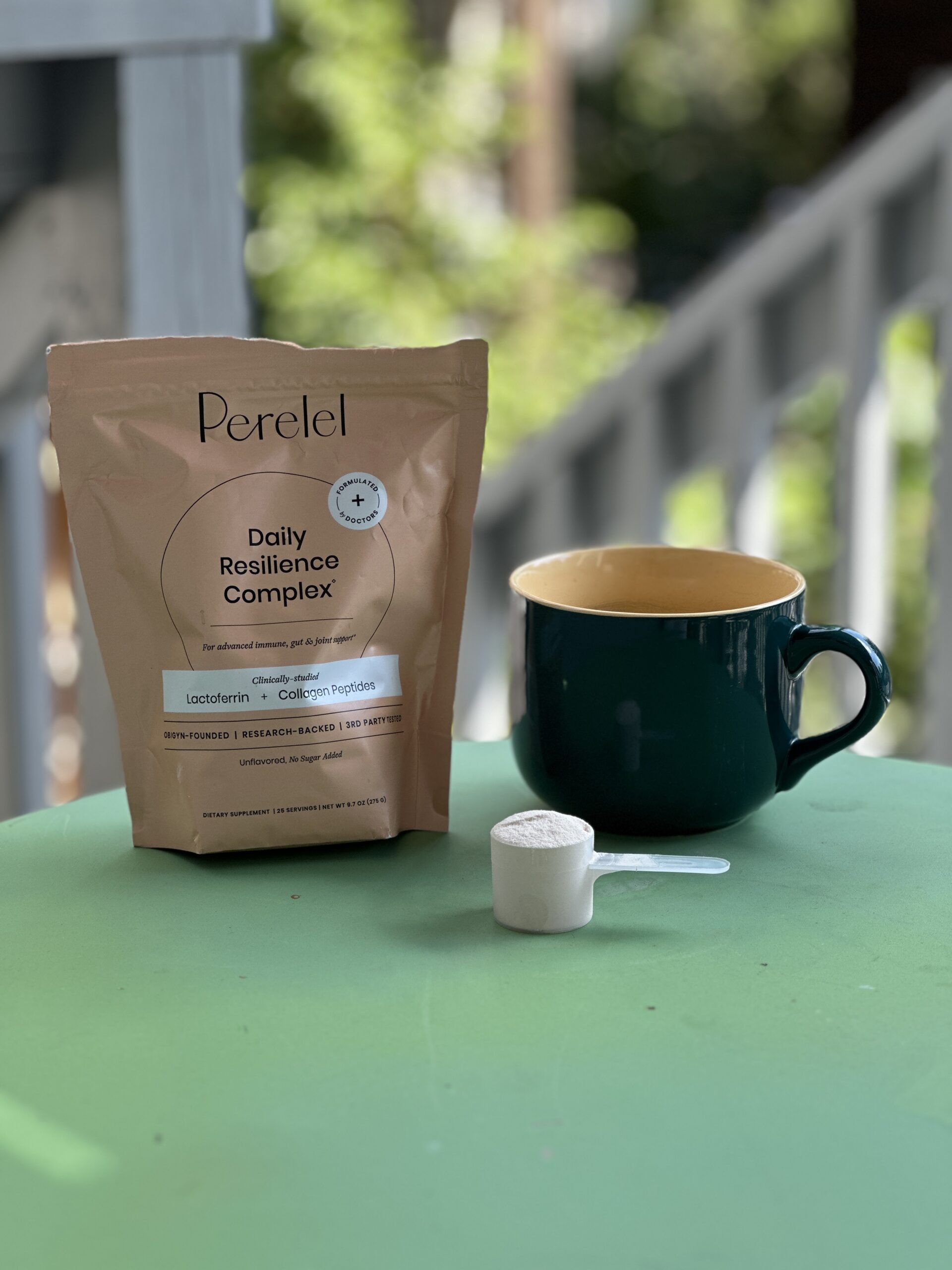 A bag of Peretel Daily Resilience Complex, a green mug, and a scoop of white powder on a green table with a blurred outdoor background.
