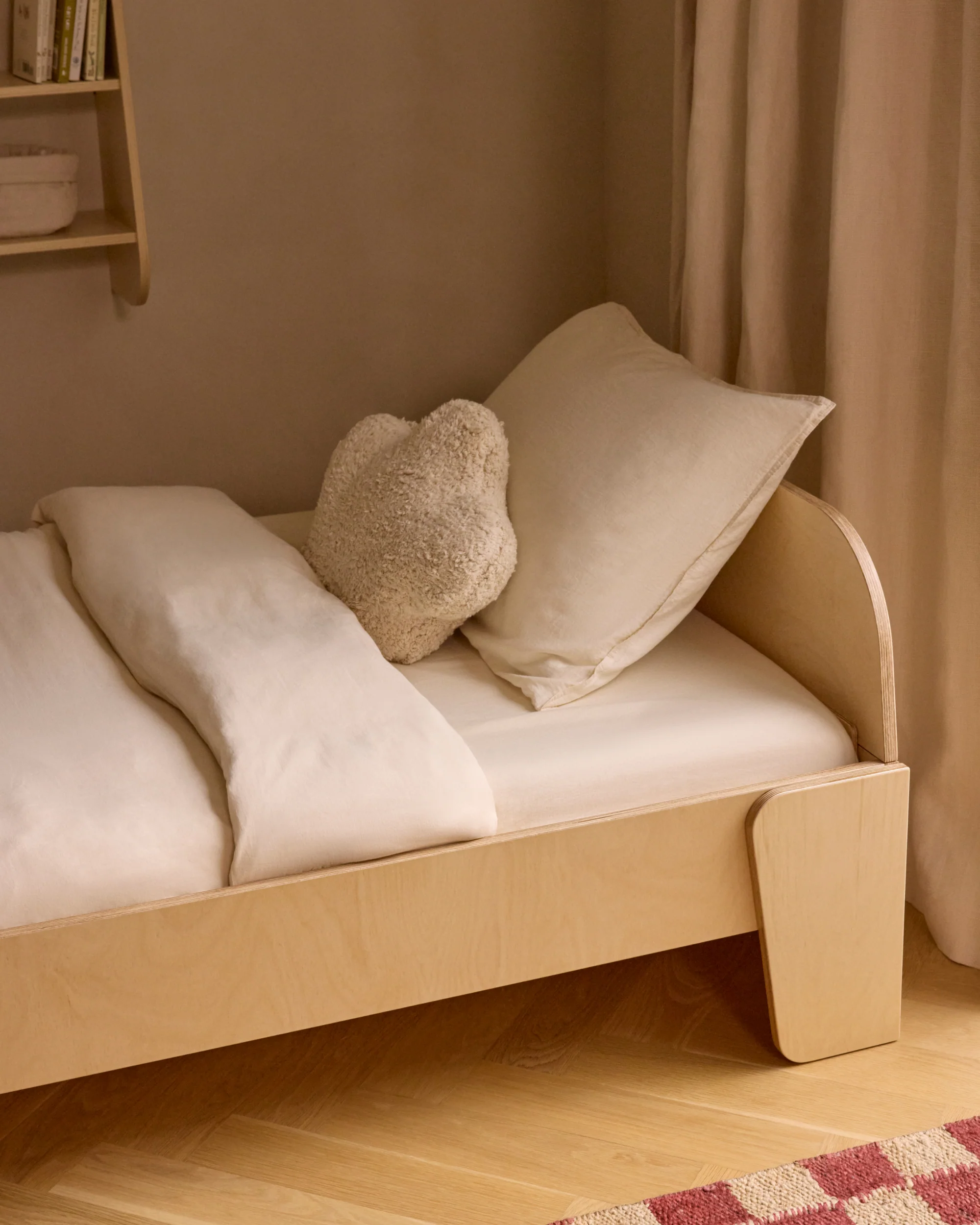 A minimalist wooden bed with white bedding, a heart-shaped pillow, and a cream curtain in a softly lit room.
