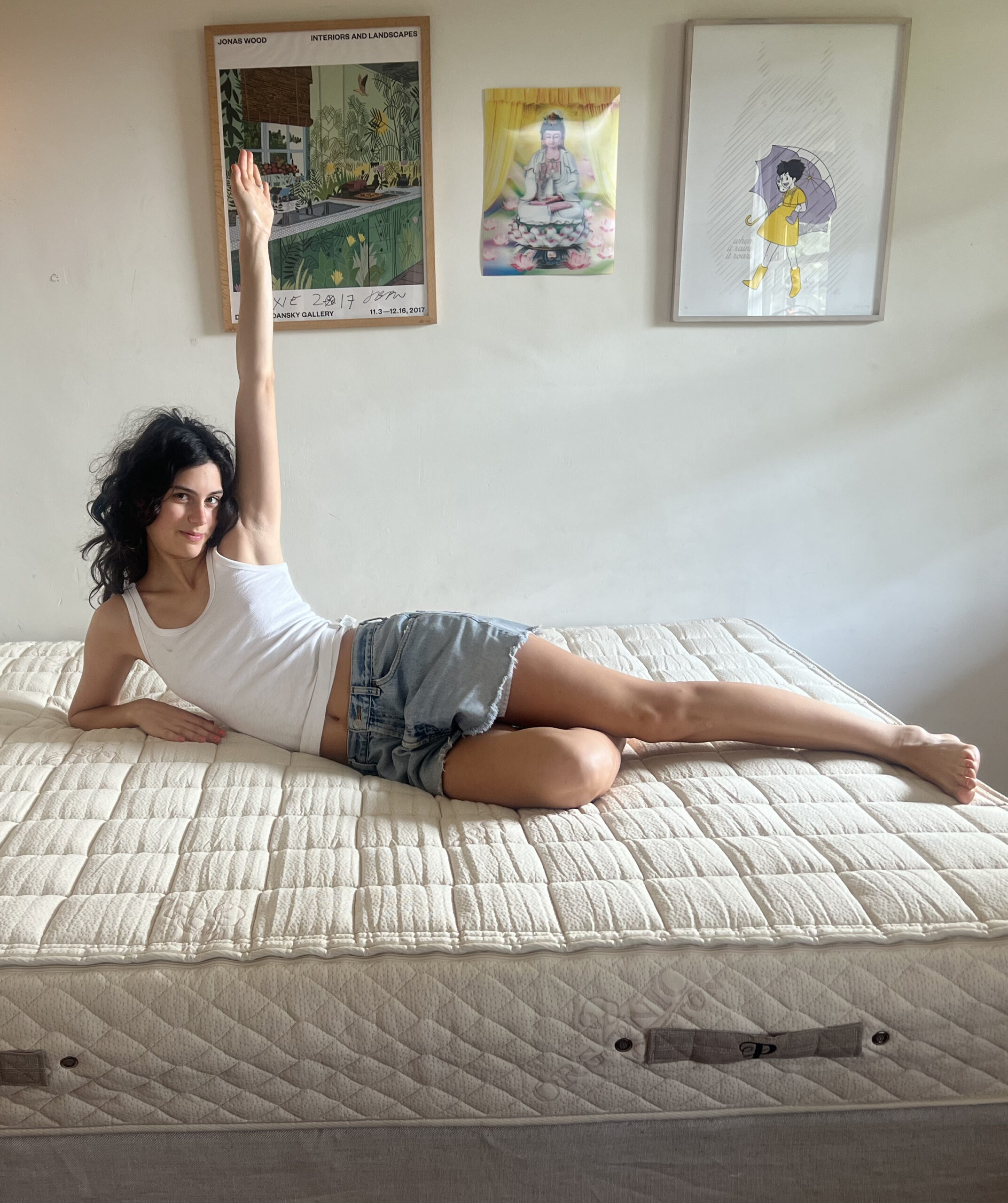 A woman in a white tank top and denim shorts lies on a mattress, propped up on one arm with the other arm raised, in a room with framed artwork on the wall.