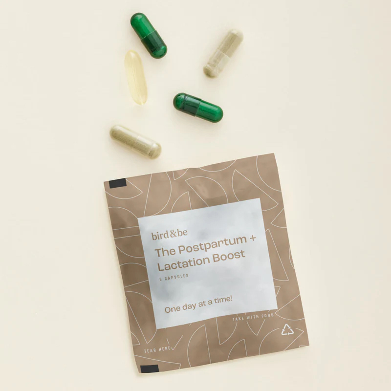 A packet labeled “The Postpartum + Lactation Boost” by bird&be is placed on a light surface next to five assorted capsules and pills.