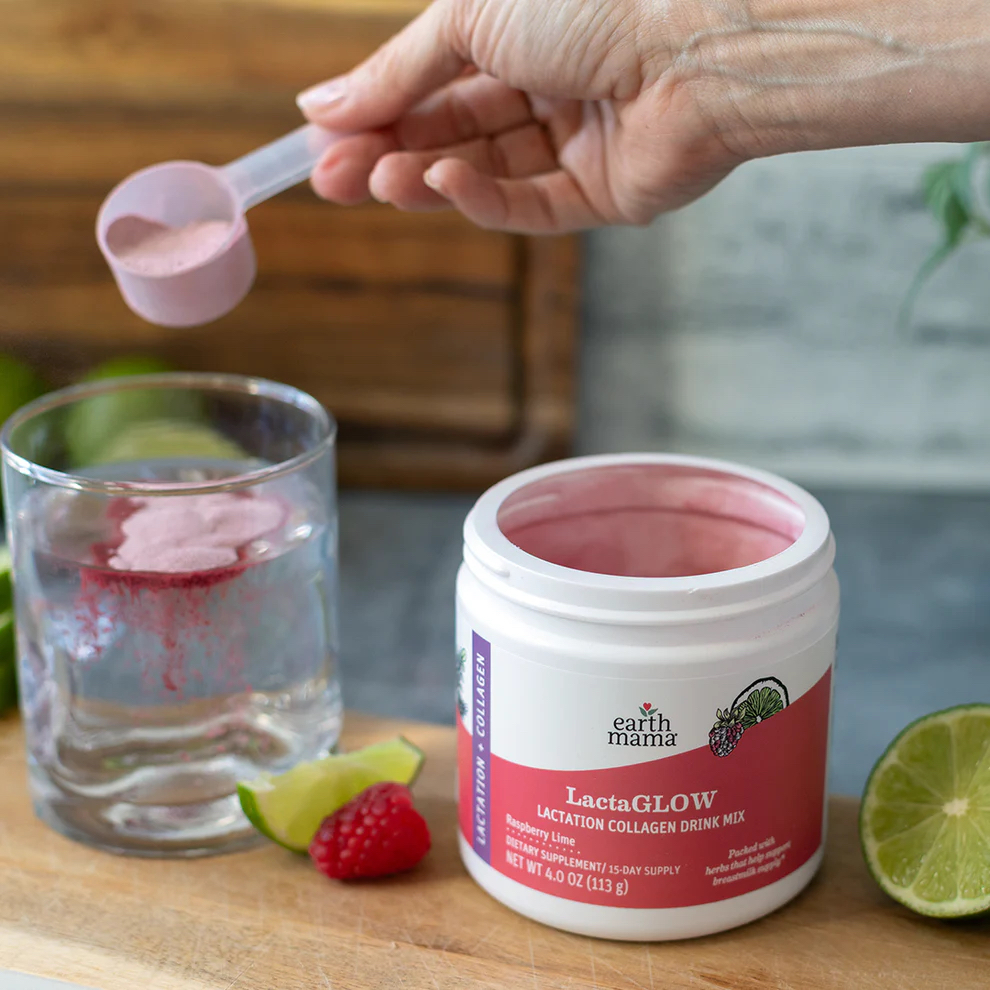 A hand pours a scoop of pink LactaGLOW collagen drink mix into a glass of water next to an open container, lime, and raspberries on a wooden surface.