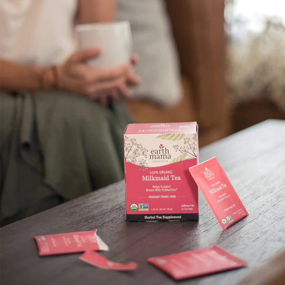 A box of Earth Mama Organic Milkmaid Tea with several tea packets on a table; a person holding a mug is blurred in the background.