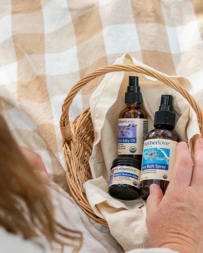 A hand holds a wicker basket containing bottles of Motherlove Sitz Bath Spray and Motherlove Birth & Baby Oil, on a beige and white checkered cloth.