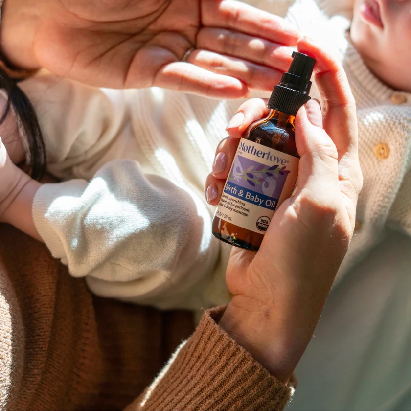 A person holds a bottle of Motherlove Birth & Baby Oil near a baby dressed in a light-colored outfit.